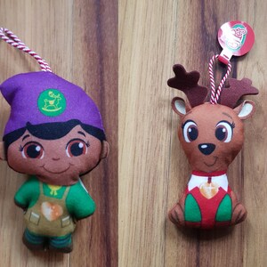 May include: Two plush Christmas ornaments, one is a purple-hatted elf with a green shirt and brown pants, the other is a brown reindeer with a red and white shirt. Both have smiling faces and are holding a heart-shaped ornament.