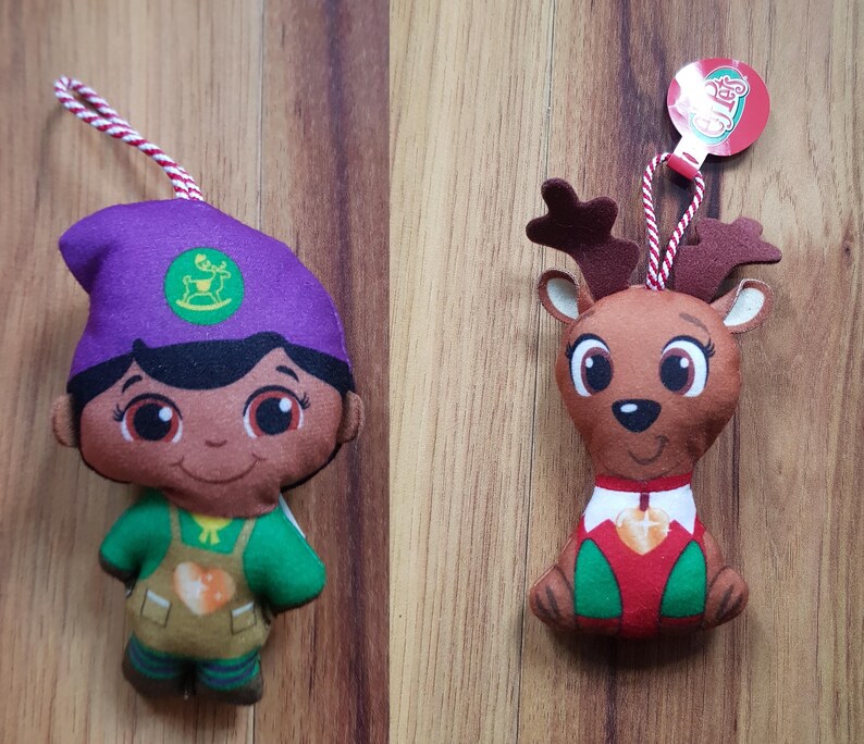 May include: Two plush Christmas ornaments, one is a purple-hatted elf with a green shirt and brown pants, the other is a brown reindeer with a red and white shirt. Both have smiling faces and are holding a heart-shaped ornament.