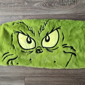 Könnte beinhalten: Ein grünes, plüschiges Kissen mit dem Gesicht des Grinch. Die Augen des Grinch sind gelb mit schwarzen Umrissen, und sein Mund ist verkniffen. Das Kissen hat ein Etikett mit dem Wort "Grinch" daran.