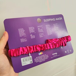 Pu&ograve; includere: Una maschera per dormire in raso rosa con un'etichetta bianca che dice "702AY-43X-ONE" e "E.A.C." La confezione &egrave; viola e ha il testo "SLEEPING MASK" e "sinsay.com".