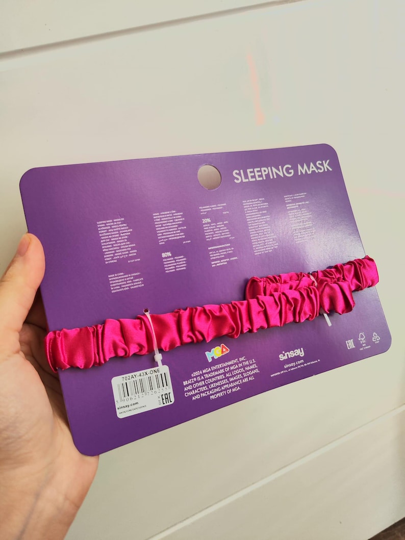 Pu&ograve; includere: Una maschera per dormire in raso rosa con un'etichetta bianca che dice "702AY-43X-ONE" e "E.A.C." La confezione &egrave; viola e ha il testo "SLEEPING MASK" e "sinsay.com".