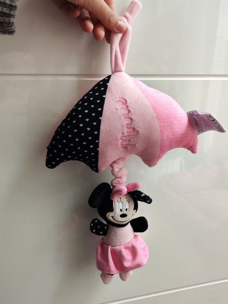 K&ouml;nnte beinhalten: Rosa-schwarzes Babyspielzeug mit einer Minnie Mouse Figur, die an einem rosa-schwarzen Wolkenf&ouml;rmigen Pl&uuml;schtier h&auml;ngt. Die Wolke hat wei&szlig;e Punkte und den Text "Minnie" aufgestickt.