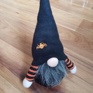 Op de afbeelding: Een Halloween-kabouter met een zwarte hoed met een oranje spinnendesign. De kabouter heeft een grijze baard, een ronde neus en armen met oranje en zwarte strepen. De armen van de kabouter hebben witte uiteinden.