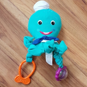 Può includere: Un giocattolo per bambini a forma di polpo in peluche turchese con un cappello da chef bianco, un papillon blu e un anello per la dentizione arancione. Il giocattolo ha una faccia sorridente con occhi neri e bocca rossa.