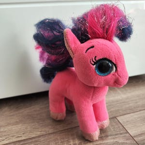 Op de afbeelding: Roze pluche pony speelgoed met een blauwe en roze manen en staart. De pony heeft grote blauwe ogen en een glinsterende afwerking.