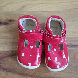 Pu&ograve; includere: Un paio di scarpe rosse in vernice per bambini piccoli con pois bianchi e orecchie di Topolino. Le scarpe hanno cinturini in velcro e all'interno c'&egrave; scritto "Disney Clarks".