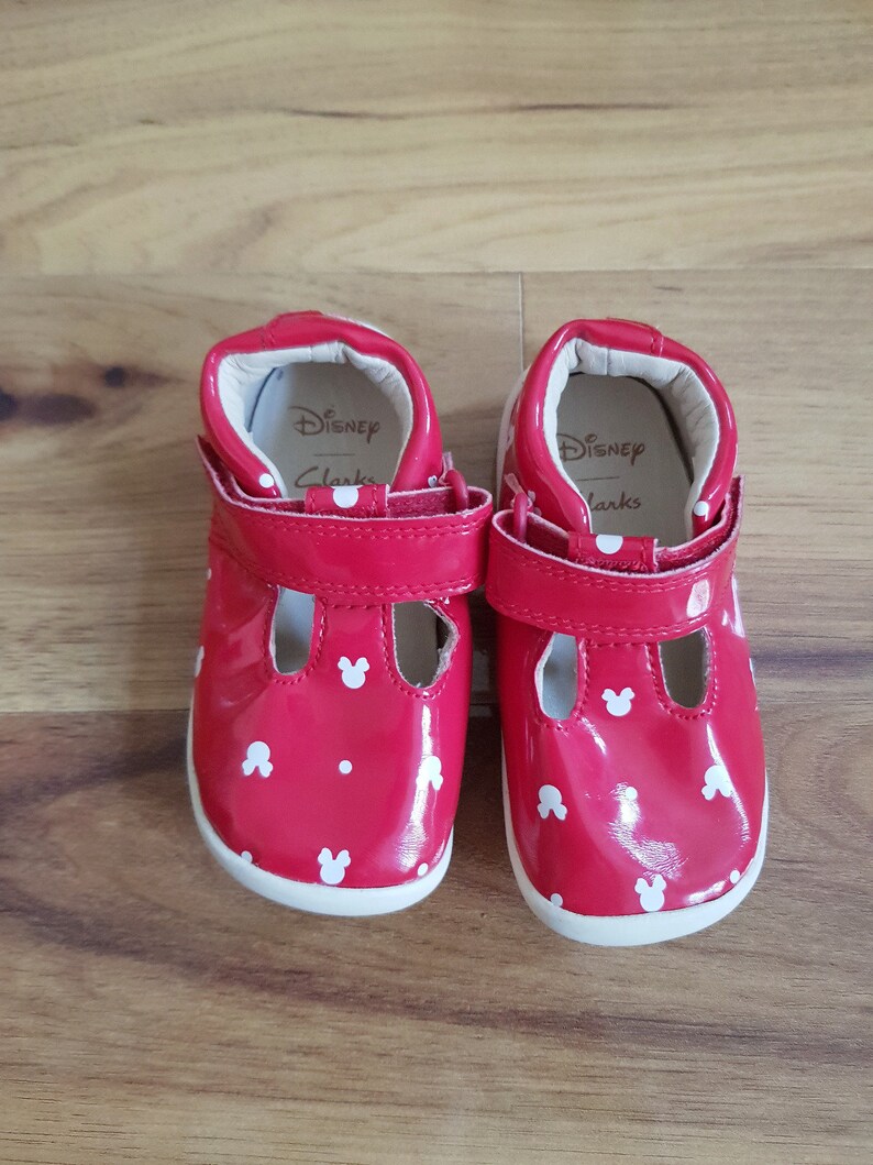 Pu&ograve; includere: Un paio di scarpe rosse in vernice per bambini piccoli con pois bianchi e orecchie di Topolino. Le scarpe hanno cinturini in velcro e all'interno c'&egrave; scritto "Disney Clarks".