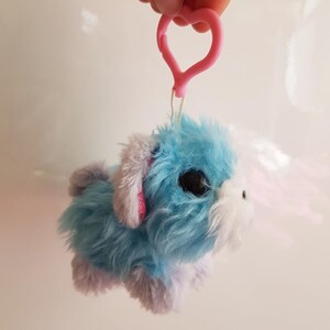 May include: A plush toy dog with blue and purple fur, black eyes, and a white muzzle. It has pink accents on its ears and a pink heart-shaped clip. The toy is designed to be a keychain or a decorative item.