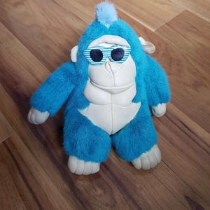 Peut inclure: Une peluche gorille bleu avec un visage blanc et des détails blancs. Le gorille porte des lunettes de soleil à rayures bleues et blanches. Il y a une touffe de fourrure bleue sur sa tête, posé sur une surface en bois.