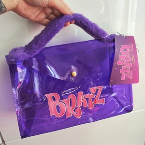 Puede incluir: Una bolsa de plástico transparente de color morado con un asa de pelo morado. La bolsa tiene un logotipo "Bratz" rosa con un halo sobre la "z".