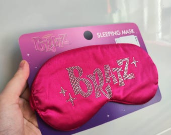 Può includere: Maschera per dormire in raso rosa con la parola "Bratz" in strass. La maschera è confezionata su una carta viola con le parole "Sleeping Mask" stampate su di essa.