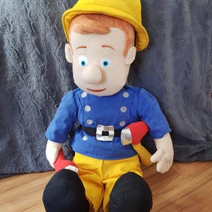 Op de afbeelding: Een pluche pop van een personage met een gele brandweerhelm met een rood embleem, een blauw jasje met zilveren knopen en een zwarte riem, een gele broek en zwarte laarzen. De pop heeft een lichte huid, blauwe ogen en oranje haar.