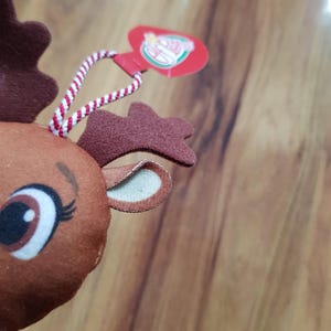 May include: Close-up of a brown reindeer plush toy with a red and white striped string attached to a red heart-shaped tag that says "The Elf on the Shelf".