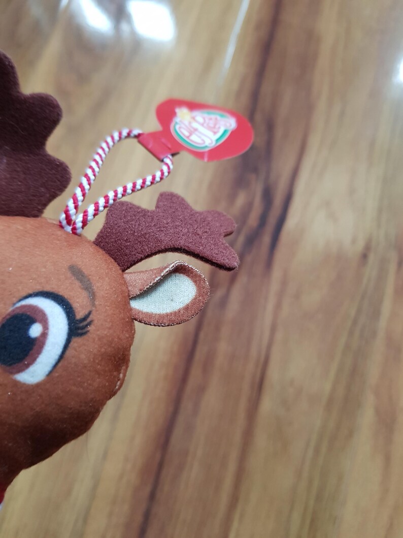 May include: Close-up of a brown reindeer plush toy with a red and white striped string attached to a red heart-shaped tag that says "The Elf on the Shelf".