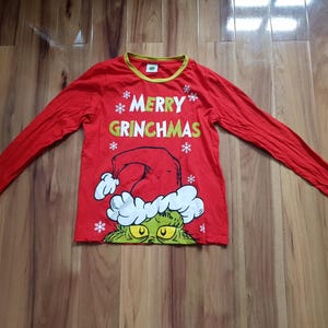 May include: Red long-sleeved shirt with a yellow neckline. The shirt features the text "MERRY GRINCHMAS" in white and green, along with snowflakes and a graphic of the Grinch wearing a Santa hat. A festive holiday-themed top.