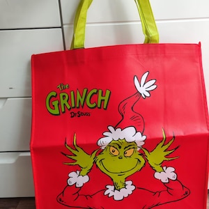 Pode incluir: Bolsa tote vermelha com o personagem Grinch de Dr. Seuss. O Grinch é verde com um chapéu e casaco vermelhos. A bolsa tem alças verdes e as palavras "The Grinch" em verde.