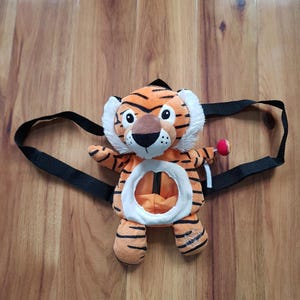 Op de afbeelding: Oranje en zwarte rugzak in de vorm van een tijger met zwarte bandjes. De pluche rugzak heeft een witte buik en een kleine rode lolly. De tijger heeft witte vacht rond zijn gezicht en zwarte strepen.