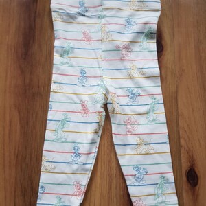 May include: White leggings with horizontal stripes in red, blue, green, and yellow. The leggings feature cartoon character illustrations. The leggings are made of a soft, stretchy material, perfect for casual wear.