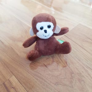 Op de afbeelding: Een kleine, bruine pluche aap met een witte gezicht, zwarte ogen en beige oren. Het knuffeldier zit op een houten oppervlak. De armen en benen van de aap zijn uitgespreid.