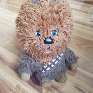 Op de afbeelding: Een bruin en zwart, pluche Chewbacca-speelgoed met blauwe ogen. Het speelgoed draagt een bruin en witte bandoleer.