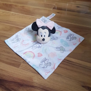 May include: A white baby comforter featuring a Minnie Mouse plush head with a pink bow. The blanket has a pattern of Minnie Mouse, rainbows, and hearts in pastel shades. The plush head is approximately 10 cm tall.