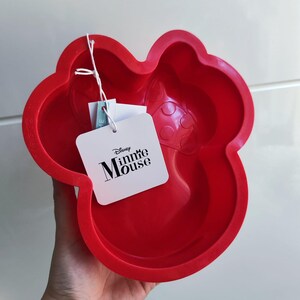 Könnte beinhalten: Eine leuchtend rote Silikon-Backform in Minnie-Maus-Form. Die Form hat ein Etikett mit dem Disney Minnie Mouse-Logo. Die Form ist zum Backen von Kuchen oder anderen Leckereien bestimmt.