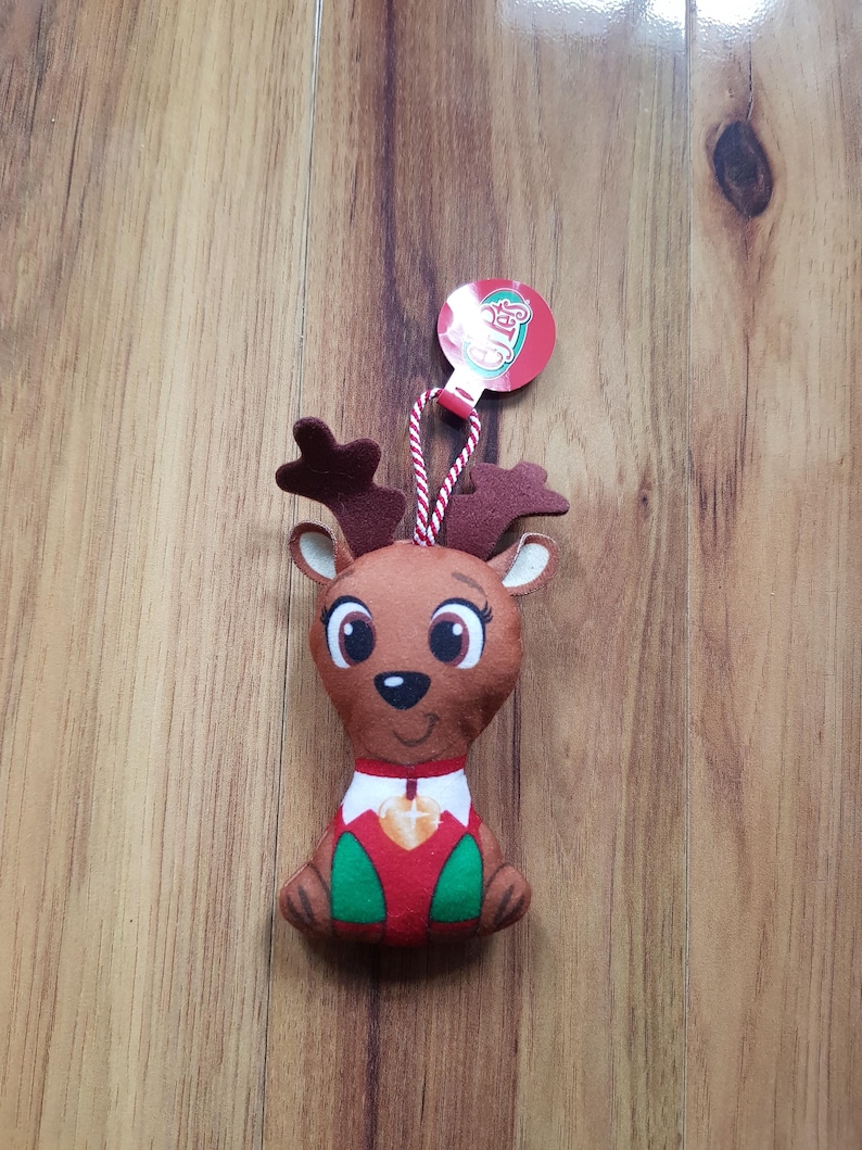 May include: A plush reindeer ornament with a red and green outfit and a brown face. The ornament has a red tag with the word "Cheers" on it.