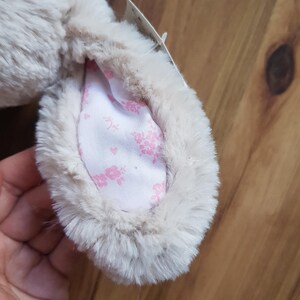May include: A close-up of a plush bunny ear. The ear is a soft, light beige colour with a fluffy texture. The inside of the ear features a white fabric with a delicate pink floral pattern. A small tag is attached.
