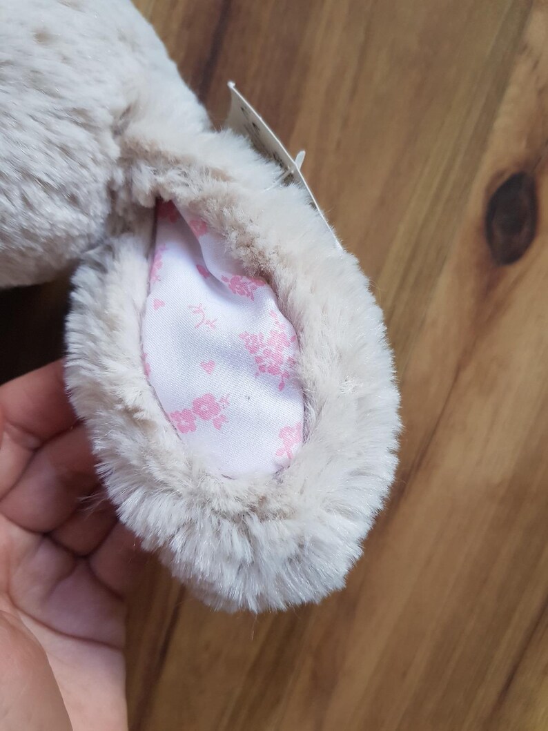 May include: A close-up of a plush bunny ear. The ear is a soft, light beige colour with a fluffy texture. The inside of the ear features a white fabric with a delicate pink floral pattern. A small tag is attached.