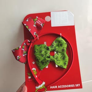 Könnte beinhalten: Ein rotes Haaraccessoires-Set mit einem Stirnband und zwei Schleifen. Das Stirnband und eine Schleife haben ein grünes Schleifendesign. Die andere Schleife ist rot mit einem grünen Muster. Das Set befindet sich auf einer roten Karte mit dem Text "HAIR ACCESSORIES SET".
