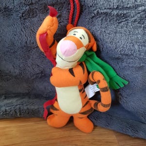 Puede incluir: Un peluche de Tigger, un personaje de tigre naranja con rayas negras, una nariz rosa y una bufanda verde. El juguete tiene un lazo rojo para colgar y está hecho de tela suave. El juguete es un personaje de la serie Winnie the Pooh.
