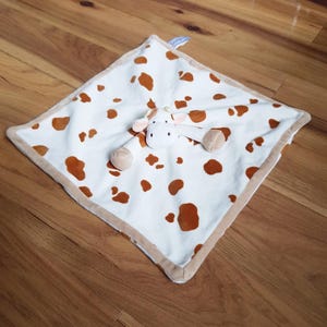 Può includere: Una coperta di sicurezza per bambini con un motivo a mucca. La coperta bianca ha macchie marroni e un bordo marrone. Include una testa e delle braccia di mucca di peluche. Realizzata in materiale morbido, ideale per i bambini.