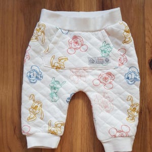 May include: White quilted baby trousers with colourful cartoon character outlines. The trousers have a pocket and elastic cuffs. The characters include Mickey Mouse, Donald Duck, Goofy, and others. The trousers are made of a soft, quilted fabric.