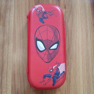 Peut inclure: Trousse rouge avec des graphismes de Spider-Man. L'étui a une fermeture éclair et une forme rectangulaire. Le design comprend le masque de Spider-Man et des poses d'action en bleu et rouge.
