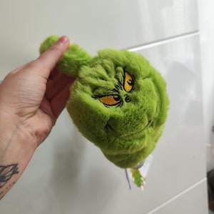 May include: A plush green Grinch head with embroidered yellow eyes and a stitched mouth. The Grinch's face is made of soft, textured fabric. The Grinch is a character from the Dr. Seuss book, "How the Grinch Stole Christmas!"