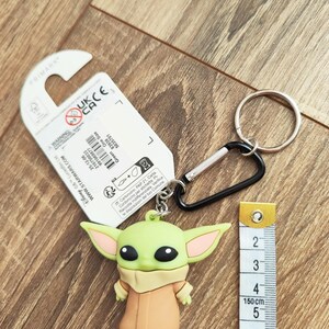 Może przedstawiać: Zielony i beżowy brelok Baby Yoda ze srebrnym k&oacute;łkiem i czarnym karabińczykiem. Brelok ma około 5 cm wysokości. Obok breloka znajduje się miarka.