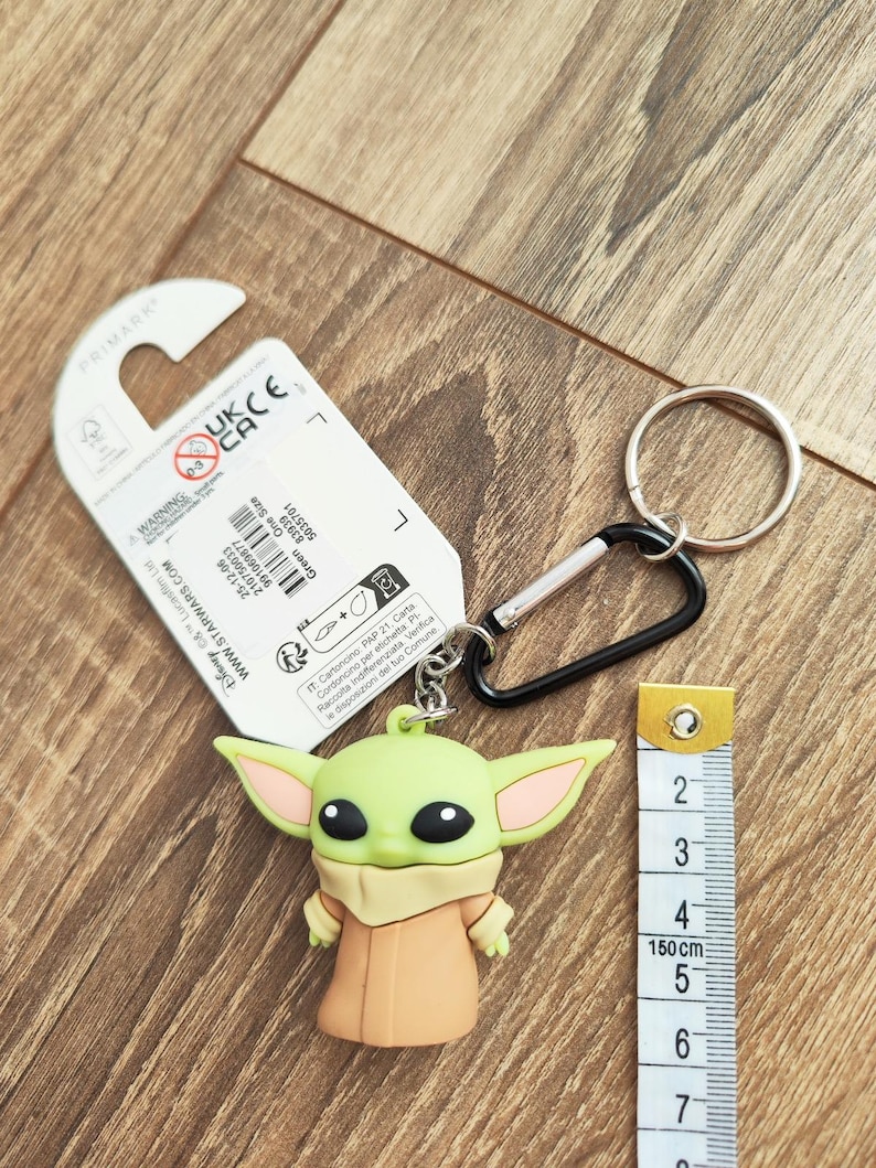 Może przedstawiać: Zielony i beżowy brelok Baby Yoda ze srebrnym k&oacute;łkiem i czarnym karabińczykiem. Brelok ma około 5 cm wysokości. Obok breloka znajduje się miarka.