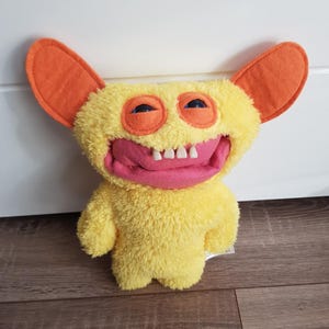 Op de afbeelding: Een gele monsterknuffel met oranje oren, blauwe ogen en een roze mond met witte tanden. Het speelgoed heeft een fuzzy textuur en een vriendelijk, cartoonachtig uiterlijk. Het is een zacht speelgoed.