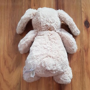 May include: A beige plush bunny toy with long, floppy ears. The soft toy is lying on a wooden surface. The bunny has a fluffy texture and a small, round tail.