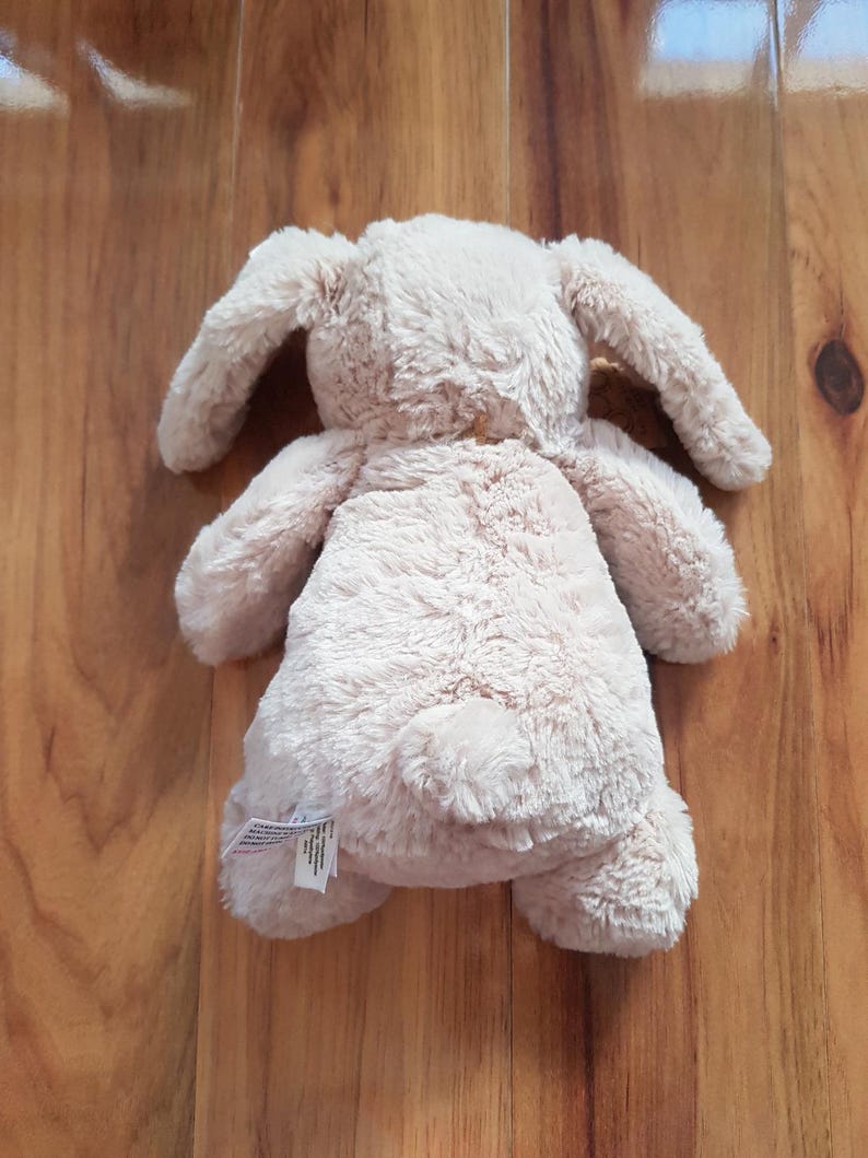 May include: A beige plush bunny toy with long, floppy ears. The soft toy is lying on a wooden surface. The bunny has a fluffy texture and a small, round tail.