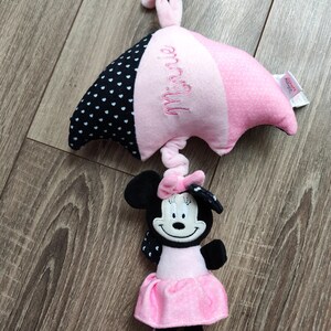 K&ouml;nnte beinhalten: Ein rosa-schwarzer Tupfen-Schirm mit dem Namen "Minnie" bestickt. Ein Pl&uuml;sch-Minnie-Maus-Spielzeug ist an der Unterseite des Schirms befestigt.