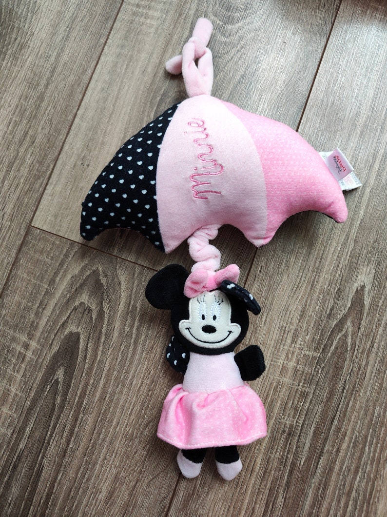 K&ouml;nnte beinhalten: Ein rosa-schwarzer Tupfen-Schirm mit dem Namen "Minnie" bestickt. Ein Pl&uuml;sch-Minnie-Maus-Spielzeug ist an der Unterseite des Schirms befestigt.