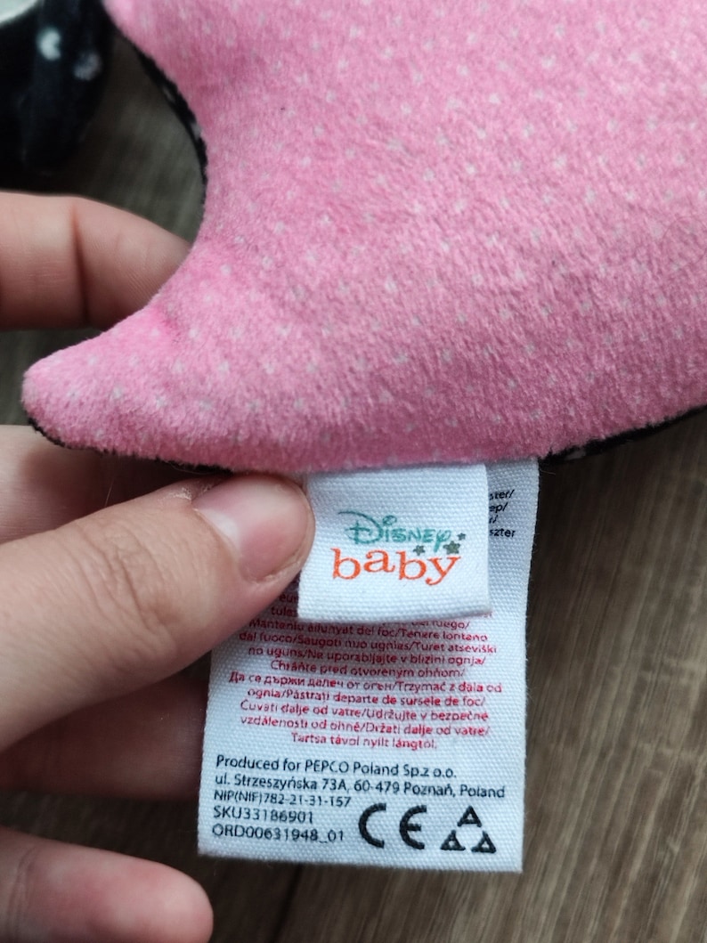 K&ouml;nnte beinhalten: Rosa-schwarzes Pl&uuml;schtier mit einem Disney Baby-Etikett. Das Etikett enth&auml;lt Pflegehinweise in mehreren Sprachen, darunter "Von Feuer fernhalten".
