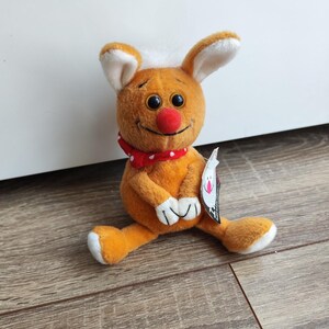 以下が含まれることがあります： 茶色の体、白い耳、赤い鼻のぬいぐるみウサギ。ウサギは赤と白のドット柄のスカーフをしています。おもちゃは木製の床の上に座っています。