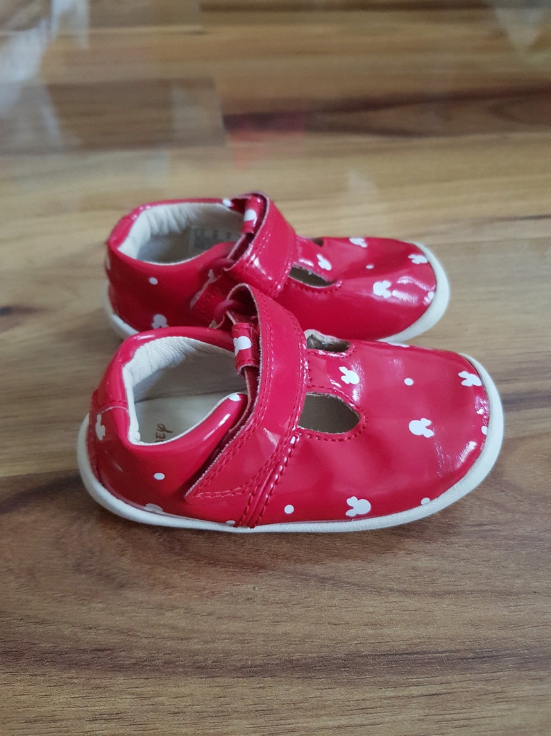 Pu&ograve; includere: Un paio di scarpine da beb&eacute; rosse con pois bianchi e orecchie di coniglio. Le scarpe hanno una chiusura a velcro.