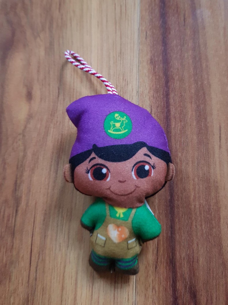 May include: Purple and green plush toy with a brown face, wearing a green shirt and brown pants. The toy has a green circle on its hat with a white reindeer inside.