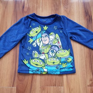 Puede incluir: Una camiseta azul de manga larga con un estampado gráfico de Buzz Lightyear y cuatro alienígenas verdes de tres ojos de Toy Story. El diseño incluye pequeños detalles de estrellas y los personajes están situados en el centro de la parte frontal.