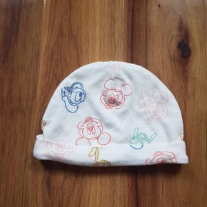 Puede incluir: Gorro de bebé blanco con contornos coloridos de personajes de dibujos animados. El gorro tiene un dobladillo y está adornado con Mickey Mouse, Donald Duck y Minnie Mouse. Fabricado con un material suave.