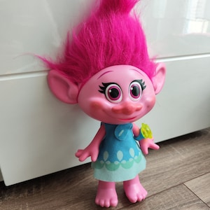 Puede incluir: Muñeca Trolls rosa y azul con pelo largo rosa, un vestido azul con detalles verdes y una pulsera verde y amarilla.