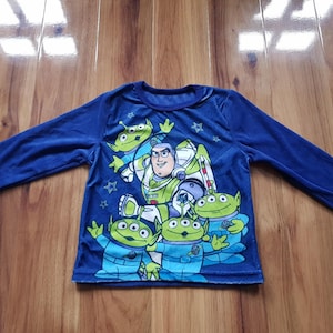 Könnte beinhalten: Ein langärmeliges, blaues Shirt mit einem Aufdruck von Buzz Lightyear und den kleinen grünen Männchen aus Toy Story. Das Shirt hat einen Rundhalsausschnitt und ist aus einem weichen Material gefertigt. Das Design umfasst Sterne und andere Charakterdetails.